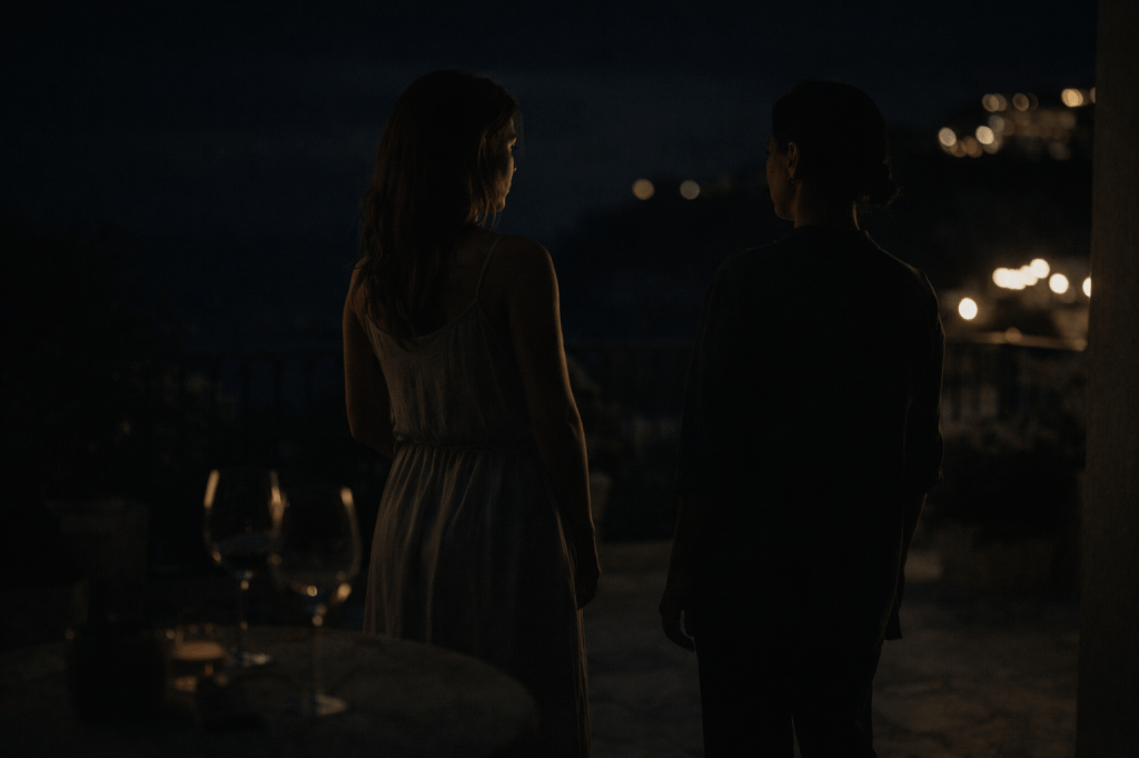 LUMIVORE V1 — PART I, IMAGE 6
“TERRACE AFTER THE NOISE”
(Night Threshold / Transition Image)
PROMPT
A horizontal, cinematic night-time photograph set on the terrace of Villa Fiorella on the Amalfi Coast, rendered in restrained A24-style realism with subtle film grain, cool moonlight tones, and deep, natural shadows.
The terrace is mostly empty after a gathering:
a small table with two half-finished wine glasses
faint condensation on the glass catching minimal light
low lanterns or string lights glowing dimly in the background, out of focus
stone flooring and terrace railings visible
the sea implied only through darkness and moving air, not scenery
The environment feels hushed, transitional, and slightly exposed — as if the party has ended but the night has not.
SUBJECT COMPOSITION
Isadora Quinn and Salomé stand near the edge of the terrace, side by side but not touching, separated by a narrow, intentional strip of space.
They are framed in three-quarter profile or from behind, partially obscured by architectural elements such as railings, columns, or shadow. Neither woman is centered in the frame.
Isadora Quinn
early–mid 30s
warm, expressive face with subtle under-eye fatigue
posture slightly open but uncertain, weight shifted unevenly
hair softly styled, catching warmer highlights from ambient light
clothing lighter in tone than Salomé’s, subtly reflecting light
expression suggests hesitation, emotional openness, a thought forming
Salomé
late 30s to early 40s
sharp, composed features
posture upright, controlled, economical
darker clothing that absorbs light rather than reflecting it
hands relaxed and still, no expressive gesture
expression unreadable, gaze angled outward or downward, not toward Isadora
They are aware of each other without engaging.
LIGHTING & TONE
cooler overall temperature than previous rooftop scenes
moonlight and distant terrace lighting edge their silhouettes
faces partially readable but not fully illuminated
no romantic glow or stylization
The image should feel like:
a pause before movement, not the movement itself
CAMERA & FRAMING
observational distance
asymmetrical composition
foreground elements (railing, glassware, shadow) intrude slightly
no direct eye contact with camera
the women are present, not posed
NEGATIVE PROMPTS (STRICT)
no walking yet
no hand-holding
no smiles
no overt romance
no scenic postcard framing
no glamour lighting
no centered composition
This image exists to mark the moment before “just for air.”
Nothing is resolved.
Nothing is claimed.