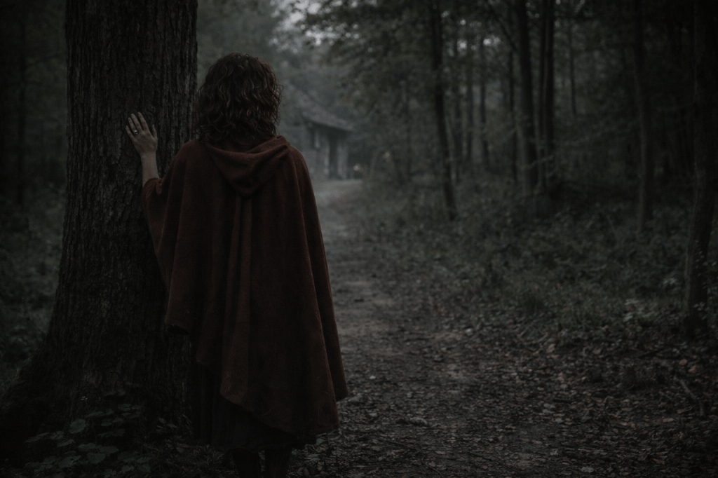 IMAGE 4 — AFTERMATH / STILLNESS

“The woods are listening again.”
Part II Cover Image

FINAL LOCKED IMAGE PROMPT (ARCHIVAL)

A horizontal, photorealistic cinematic still set at the edge of an ancient French forest just beyond a stone cottage.

The immediate aftermath of violence is present, but only indirectly.

The cottage itself is not the subject. It appears only in suggestion at the edge of the frame or softly out of focus in the background — a darkened doorway, a damaged shutter, subtle signs of disruption without spectacle. No bodies. No blood. No visible aftermath staged for display.

In the foreground stands Red Valois, alone.

She is a young French woman in her late 20s with unruly chestnut-brown curls, worn loose and natural, slightly damp and disordered from exertion. Her hair frames her face unevenly, with stray curls resting against her cheeks and neck — realistic, unstyled, and consistent with physical movement rather than presentation.

Her skin is olive-toned. Her features are grounded and natural, not idealized. Her expression is calm, serious, and inward — not victorious, not shaken, but settled.

She stands near an old tree at the forest’s edge — oak or beech — its bark rough, scarred, and familiar. One hand rests flat against the trunk, fingers spread, touching it instinctively rather than ceremonially, as if grounding herself after violence.

Her posture is upright but quiet. Balanced. Still.

She wears her simple weathered red wool cloak, draped naturally over her shoulders. The cloak is slightly disordered: creased from movement, the hem darkened with dirt. It no longer reads as symbolic or heroic — it is simply clothing again. Beneath it, a loose off-white linen blouse and a dark wool skirt, practical and worn.

The forest is calm but not pristine.

Leaves lie disturbed near the path. Underbrush is bent or broken where something passed through. The ground shows scuffed earth and displaced foliage — signs of interruption already beginning to settle.

Lighting is soft, natural, and cool — dawn or just-after. No golden triumphal light. No glow. No stylization. The forest feels watchful, not welcoming.

There is no Fenris visible.
No body. No trace centered in frame.
His absence is felt through space rather than image.

The camera framing is medium-wide and observational. Red is slightly off-center. The forest occupies more visual weight than she does.

The mood is quiet, grounded, and unsettled — not because danger remains, but because power has been exercised and cannot be undone.

This is not peace.
This is equilibrium restored at a cost.

🔒 REFUSAL LOCKS (ABSOLUTE)

No heroic group tableau

No bodies or remains

No blood or gore

No triumphal or sentimental framing

No glowing magic or symbolic light

No centered or dominant Fenris presence

No overt emotional release from Red

No “happily ever after” tone