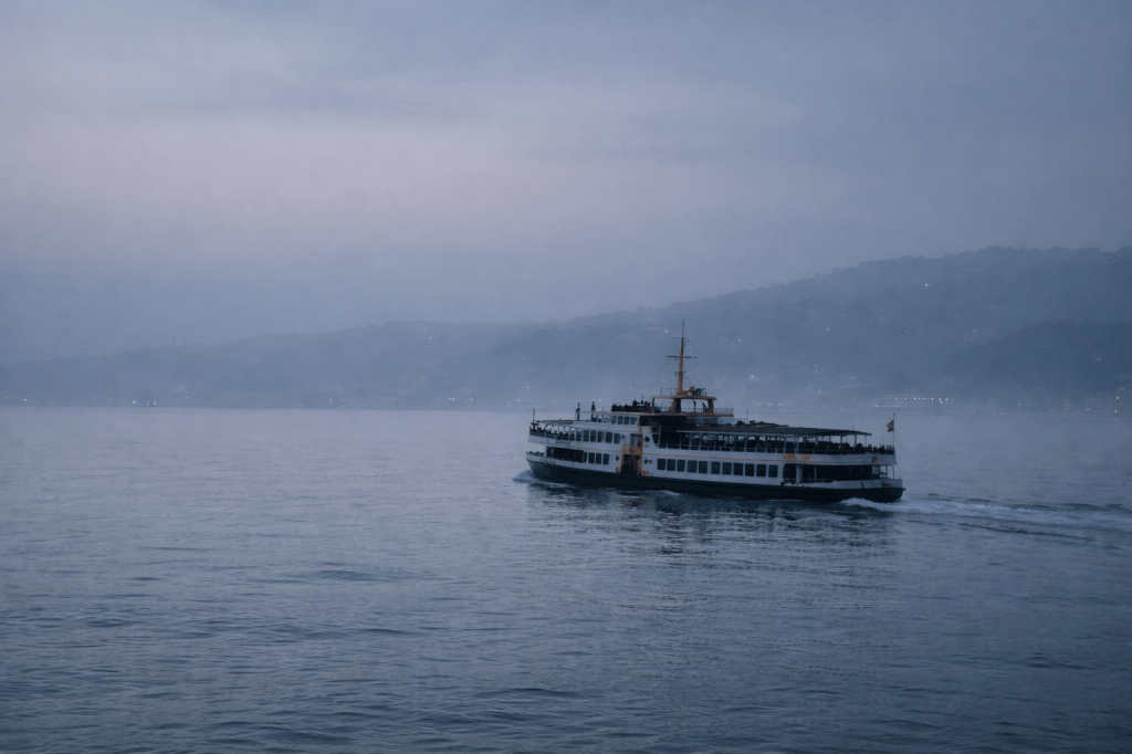 LUMIVORE V1 — ARCHIVAL STILL
“Bosphorus Crossing, Before Morning”

PROMPT (FINAL USED VERSION):

A horizontal, observational photograph of an Istanbul Bosphorus ferry in motion during very early dawn, rendered in clean, naturalistic realism.

The ferry is mid-crossing, viewed from a slight distance across calm, pale gray-blue water.
The vessel moves quietly through the strait with no visible urgency; the wake is soft and subdued, motion implied but not emphasized.

The sky is in a pre-sunrise state — cool blue-grey tones with no warmth, no golden light, and no sunrise color.
Low mist hangs over the water, gently softening edges and slightly reducing contrast.

The surrounding environment is minimal and atmospheric.
Shorelines appear only as distant, softened silhouettes, with no landmark emphasis and no architectural detail drawing focus.

Any people visible on the ferry are extremely small, indistinct, and unidentifiable; no faces in focus, no individual emphasized.

Lighting is cool, flat, and undecided — natural dawn light before the day commits to itself.
The overall mood is liminal, quiet, and transitional, conveying passage without destination.

IMAGE DISCIPLINE

Natural color only

Clean digital clarity

No film grain

No analog texture

No cinematic color grading

Slight atmospheric softness

Neutral contrast

CAMERA & COMPOSITION

Eye-level or slightly elevated viewpoint

Moderate focal length (approx. 35–50mm equivalent)

Balanced, unobtrusive framing

Emphasis on space, atmosphere, and stillness rather than action

NEGATIVE PROMPT

Avoid warm light, sunrise glow, golden-hour tones, dramatic contrast, cinematic lighting, sharp highlights, saturated color, motion blur, stylization, vignette effects, narrative staging, symbolic emphasis, or identifiable individuals.