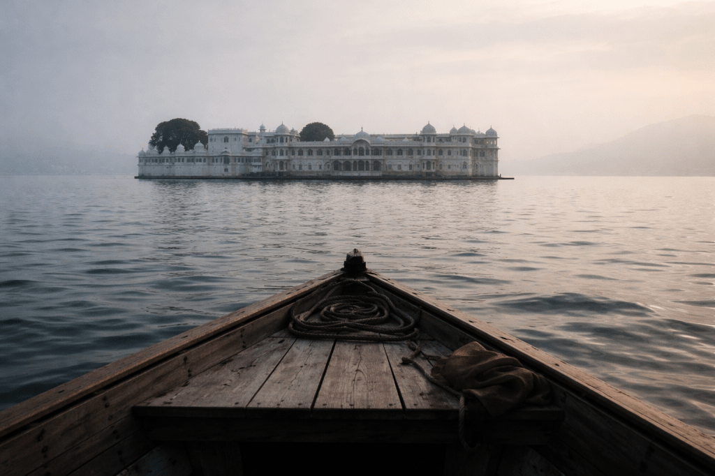 HORIZONTAL CINEMATIC IMAGE (16:9)
Prestige cinematic realism. Observational still.

A small wooden boat moves slowly across Lake Pichola toward the Taj Lake Palace in Udaipur, India. The palace is unmistakable—white marble structure rising directly from the water—yet framed without spectacle. It sits calmly at mid-distance, grounded and still, not glowing, not romanticized.

The water is muted and calm, lightly rippled, absorbing sound rather than reflecting light. The city is absent from view. No crowds. No drama.

Camera position is low, at seated eye level inside the boat, creating the feeling of approach rather than arrival. Any human figures are distant and indistinct—never the subject, never identifiable.

Lighting is early morning: soft, cool, and diffused. Pale stone, blue-grey water, faint atmospheric haze. No golden-hour warmth. No saturated color.

The composition allows breathing room around the palace. Nothing feels urgent. Nothing feels staged.

The image should feel like a deliberate retreat from noise into chosen stillness.

Location Integrity Locks (important)

The palace must read clearly as Taj Lake Palace, not a generic Indian palace

Architecture should match white marble, low domes, and lake-anchored symmetry

No signage, logos, or tourism cues

No exoticized framing

This is a lived place, not a destination.

Emotional Calibration Check

If the image feels:

calm ✔️

intentional ✔️

observational ✔️

quietly grounded in India ✔️

It’s right.

If it feels:

luxurious ❌

spectacular ❌

travel-magazine-like ❌

It’s wrong.

REFINEMENT / VARIATION PASS — MAINTAIN ALL PRIOR CONSTRAINTS

Preserve the same cinematic composition, location, and mood, but introduce subtle imperfection and lived realism.

The boat is no longer perfectly aligned with the palace—its angle is slightly off-center, suggesting gentle drift rather than precise approach. The palace sits just left or right of center in the frame, creating natural asymmetry.

The water surface shows mild irregular texture—small, uneven ripples that break perfect reflection. The lake absorbs light unevenly, with faint tonal variation rather than smooth gradients.

Morning haze is slightly inconsistent: thin patches of mist vary in density, softening parts of the palace more than others. The sky is calm but not perfectly uniform—subtle cloud diffusion breaks visual perfection.

Lighting remains soft and cool, but with minor natural variation—no painterly smoothness, no idealized gradients.

The overall effect should feel human and unplanned, as if the camera captured the moment while gently adjusting balance, not staging it.

Imperfection Intent (do not override)

Asymmetry over symmetry

Texture over polish

Drift over precision

Reality over ideal

The image should feel chosen, not perfected.

Calibration Check (post-generation)

If the image feels:

slightly less “perfect” ✔️

more lived-in ✔️

more bodily (as if sitting in the boat) ✔️

Then the refinement worked.

If it feels:

messy ❌

dramatic ❌

destabilized ❌

Then the correction went too far.