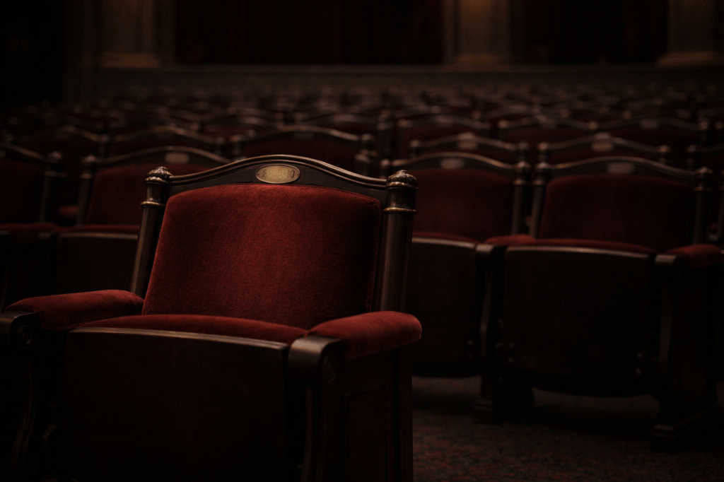 HORIZONTAL CINEMATIC IMAGE — 16:9 — OBSERVATIONAL REALISM

A quiet, empty opera house interior before a performance.

The camera is positioned at seated audience eye level, using a natural 35–50mm perspective. The framing is horizontal and slightly off-center. A single red velvet opera seat occupies the lower-left portion of the frame, partially cropped, not centered.

The seat is unoccupied. Its velvet upholstery shows subtle wear. A small brass seat number plaque is present but softly out of focus and not clearly readable.

Surrounding seats recede evenly into shadow, maintaining clarity of form without heavy blur. Depth of field remains moderate — no dramatic background separation.

Lighting is dim, neutral, and evenly distributed — standard house lighting before a performance. No warm spotlighting. No visible light sources. No glow effect.

Colors are restrained and matte: deep red velvet, dark wood, muted brass, soft black shadows.

The atmosphere is still and formal — a space waiting without emphasis or spectacle.

This image must feel observed, not composed.
No symbolism. No nostalgia. No theatrical mood.

Negative Constraints (critical)

No spotlight beams

No dramatic contrast

No shallow-focus glamour

No visible stage

No performers or audience

No illustration or painterly style