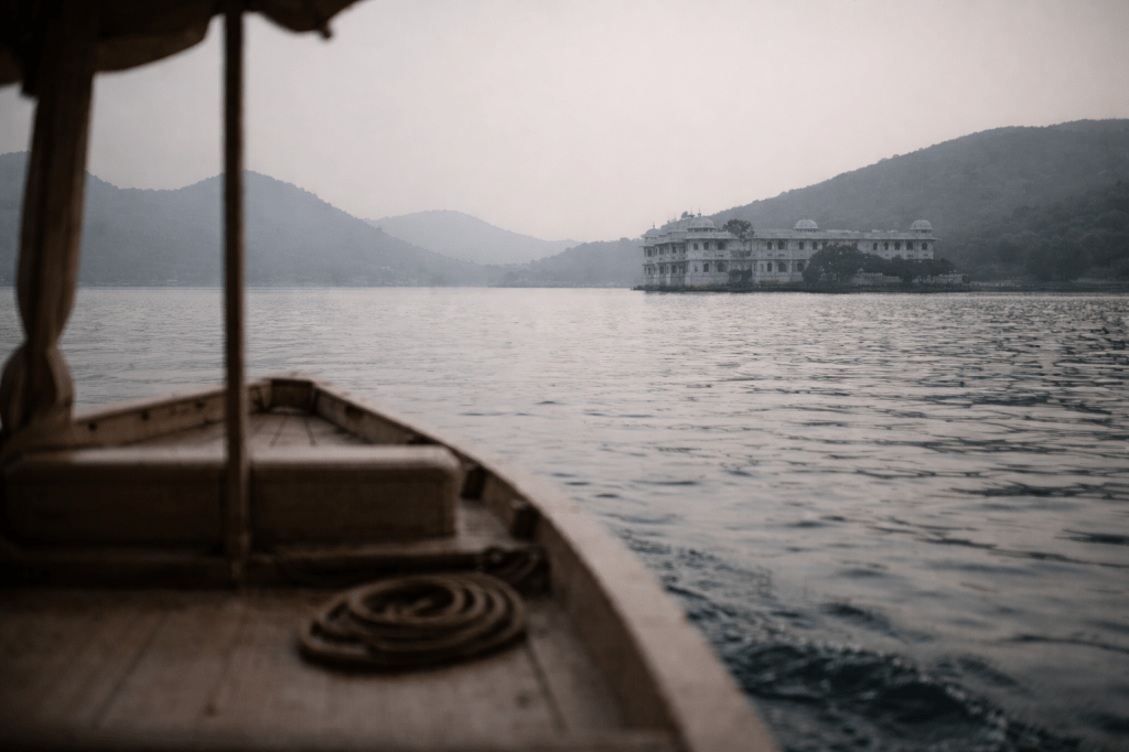 MAGE 5 — CLOSING IMAGE (REFINEMENT PASS)

Continuation / Return / Unclaimed

Maintain all prior constraints and tone.

Refinement Prompt

HORIZONTAL CINEMATIC IMAGE (16:9)
Prestige cinematic realism. Observational still.

Return to the same setting on Lake Pichola, Udaipur, India, with the Taj Lake Palace receding in the distance, softened by haze and no longer central.

Refine the human presence out of the image:

Remove visible human figures entirely or reduce them to the point of near-absence.

If a boat edge or interior is visible, emphasize material presence (wood grain, rope, edge of the hull) rather than occupants.

No faces, silhouettes, or bodies that suggest companionship or relationship.

The boat remains clearly in motion, but who is inside does not matter — or cannot be determined.

Shift emphasis forward:

Give more visual space to the water opening ahead of the boat.

Let the palace recede naturally, existing as part of the environment, not as a focal point or memory.

Light and atmosphere (unchanged, reinforced):

Neutral, diffuse light.

No warmth, no sentiment.

Mild irregular ripples on the water — steady, unremarkable.

The camera remains low and human in scale, positioned from within or just behind the boat, but without identifying presence.

The image should feel like movement continuing without narrative ownership.

Style & Restraint Locks (do not override)

No visible people

No farewell framing

No emotional light

No symbolic composition

No implication of companionship

This is motion without attachment.

Calibration Check (post-generation)

The refined image should feel:

quieter than the previous version ✔️

less narrative ✔️

more open ✔️

more anonymous ✔️

It should not feel:

lonely ❌

social ❌

resolved ❌

anticipatory ❌

Final Narrative Alignment Test

If the image feels like life continuing whether or not anyone is watching, the refinement worked.

If it invites the viewer to imagine who is there — it didn’t.